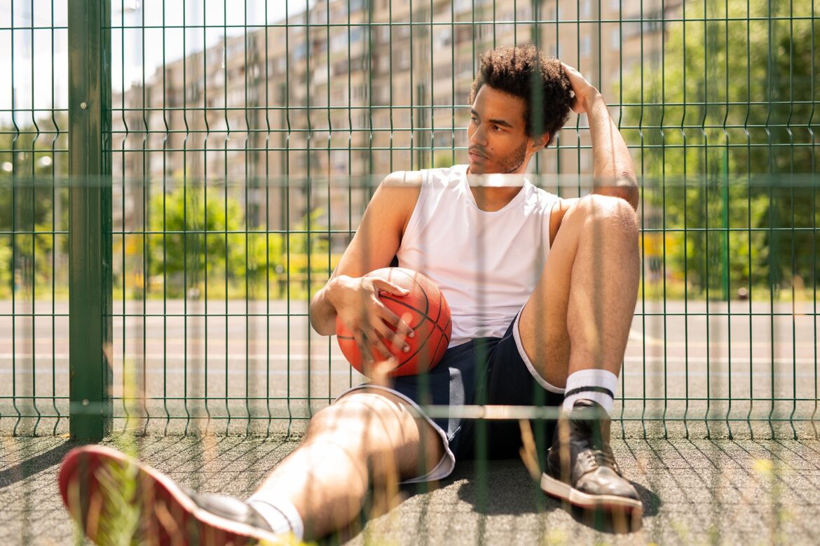 Young relaxed active man with ball sitting by fence of basketball court